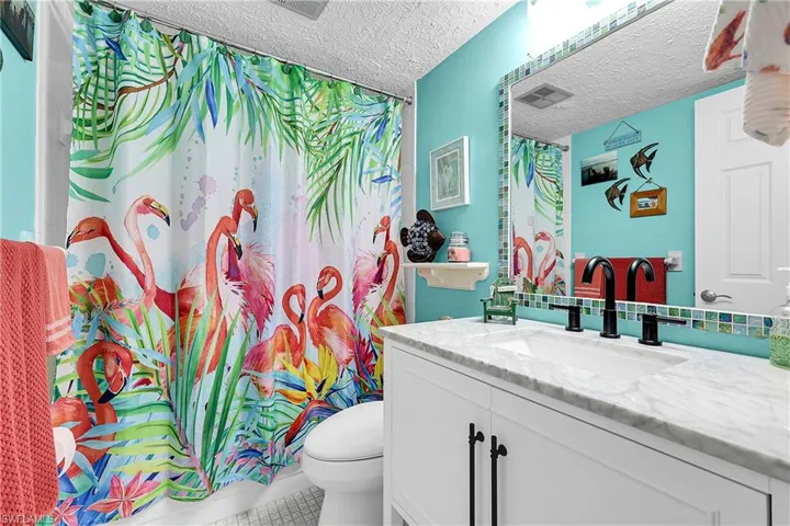 Colorful guest bathroom has updated vanity and toilet. The mirror is decorated with accent tiles. Tiled shower/tub combo is behind the flamingo curtain.