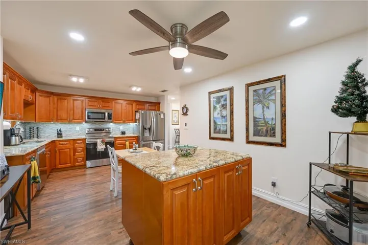 Kitchen featuring brown cabinetry, light stone counters, stainless steel appliances, a center island, and dark wood-type flooring