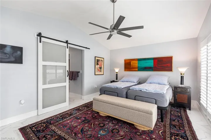 Bedroom showing porcelain tile flooring, a barn door, ceiling fan, and lofted ceiling.  Windows have wooden plantation shutters.