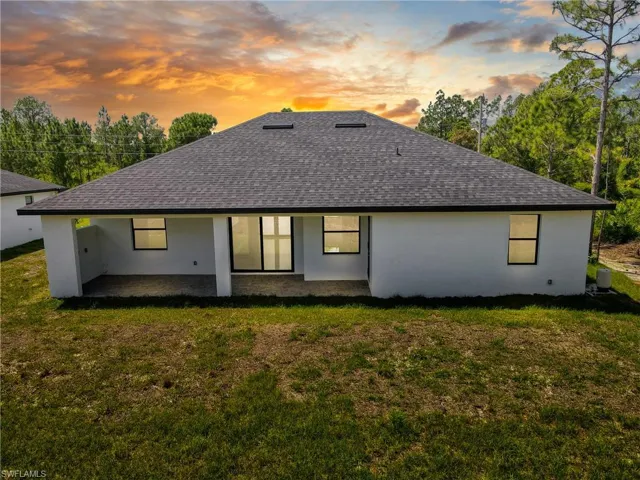 Back of house at dusk featuring roof with shingles, stucco siding, a patio, and a lawn