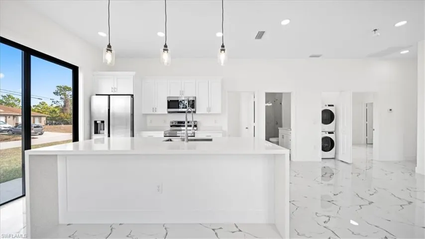 Kitchen featuring white cabinetry, stainless steel appliances, light stone counters, decorative light fixtures, and stacked washer / dryer