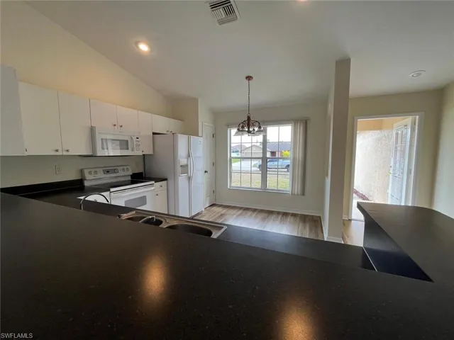 Kitchen with white appliances, hanging lights, light wood-type flooring, dark countertops, and white cabinets