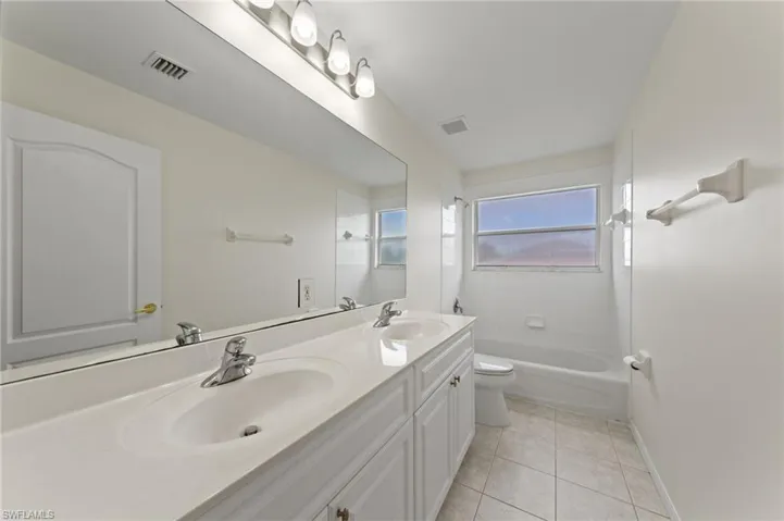 Dual vanity with integrated sinks, full-width mirror, light-toned tile flooring, and a bathtub with tile surround