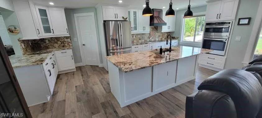 Kitchen with custom exhaust hood, appliances with stainless steel finishes, glass insert cabinets, open floor plan, and wood finished floors