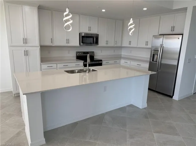 Kitchen featuring a spacious island with an undermount sink, light-toned countertops, white shaker cabinetry, stainless steel appliances, and contemporary pendant lighting