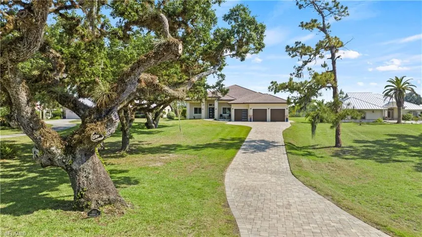 Paver driveway leading to a residence with a three-car garage and a tiled roof, set amidst a spacious lawn with mature trees and landscaping