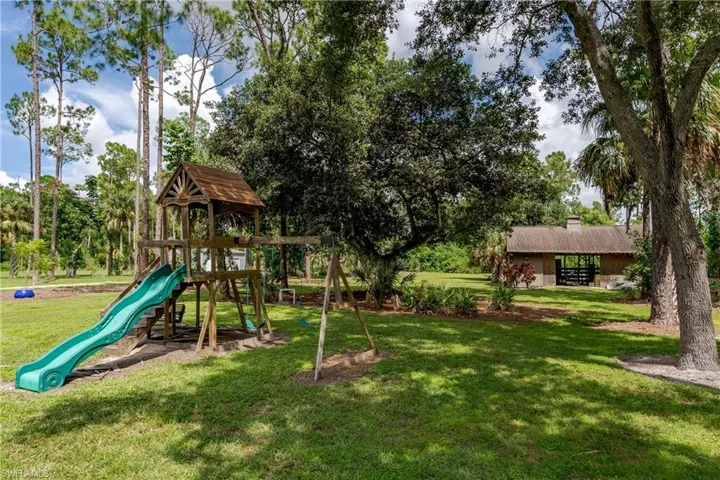 Expansive outdoor space featuring a wooden play structure with a slide and swings, mature trees, a manicured lawn, and a distant covered pavilion with a pitched roof