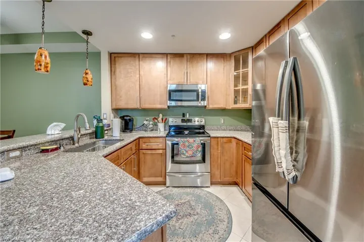 Kitchen with appliances with stainless steel finishes, glass insert cabinets, brown cabinetry, light stone countertops, and pendant lighting