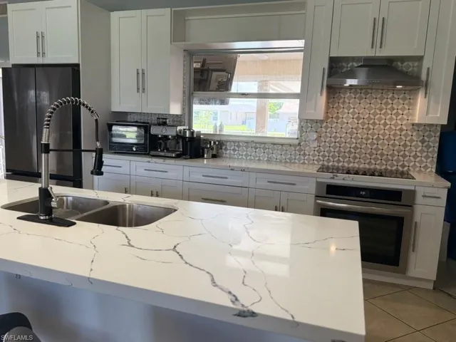 White shaker cabinetry, patterned tile backsplash, stainless steel appliances, dual basin sink, and a large island with a waterfall edge