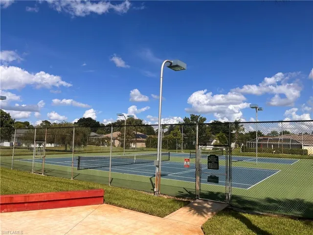 Paved tennis courts enclosed by chain-link fencing, featuring overhead lighting fixtures and an adjacent grass area