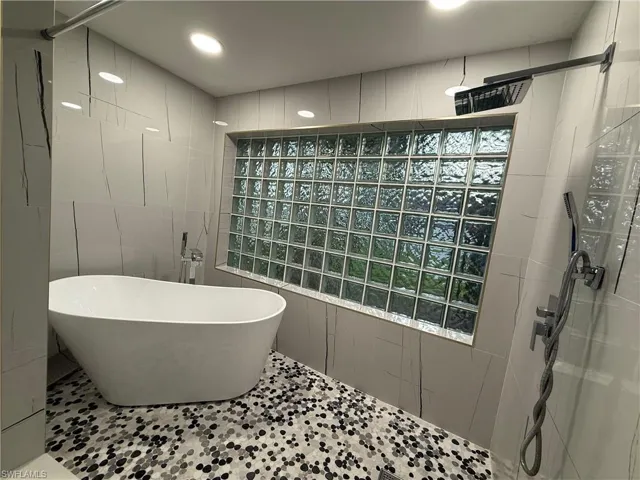 Freestanding soaking tub, mosaic tile flooring, glass block window, wall-mounted shower fixtures, and recessed ceiling lighting