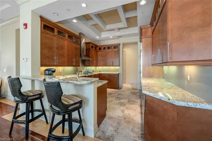 Kitchen featuring a breakfast bar area, coffered ceiling, light stone countertops, kitchen peninsula, and wall chimney exhaust hood