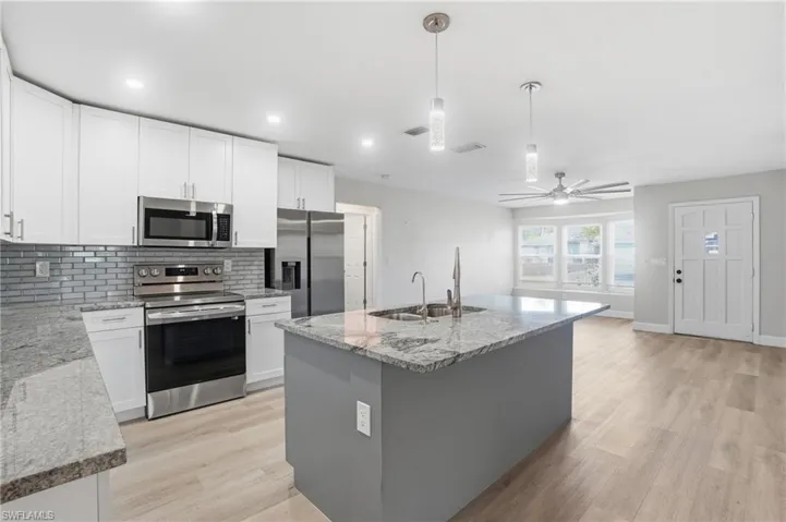 Dual tone kitchen with stainless steel appliances, a kitchen island with sink, light wood finished floors, light stone counters, and decorative light fixtures