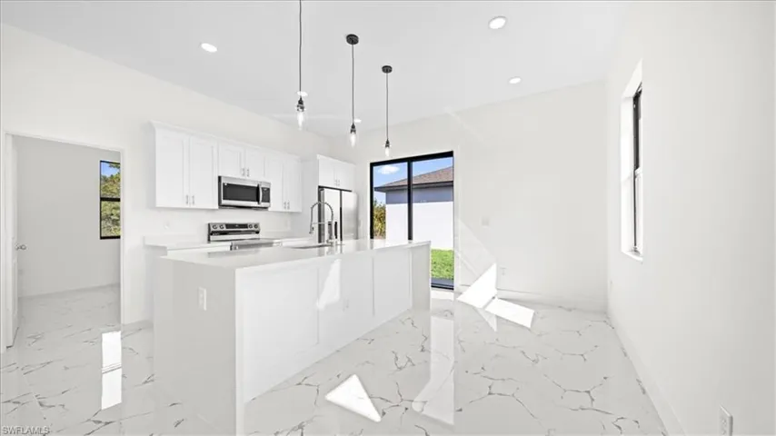Kitchen with hanging light fixtures, stainless steel appliances, white cabinetry, light marble finish flooring, and a kitchen island with sink