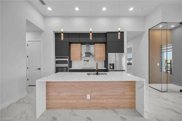 Kitchen featuring light stone counters, wall chimney exhaust hood, stainless steel appliances, hanging light fixtures, and an island with sink