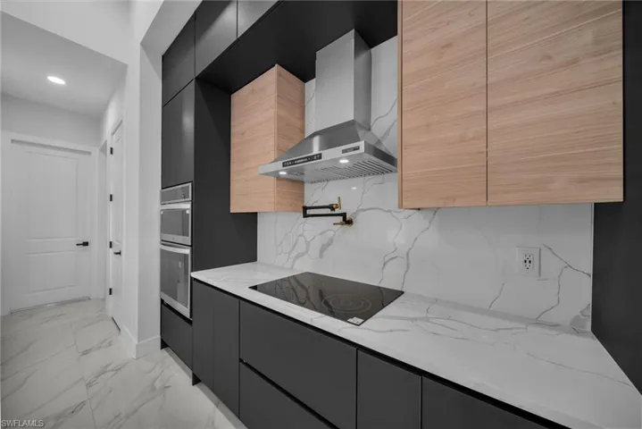 Kitchen with decorative backsplash, light stone counters, wall chimney exhaust hood, black electric cooktop, and double oven