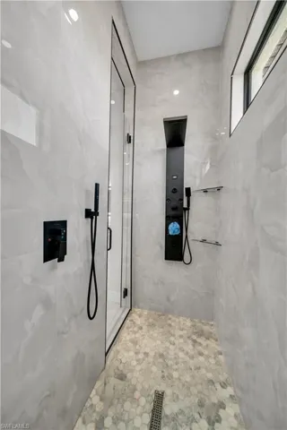 Walk-in shower featuring light-toned tile walls, hexagonal floor tiling, a multi-function shower panel with matte black fixtures, a handheld shower head, and an upper transom window