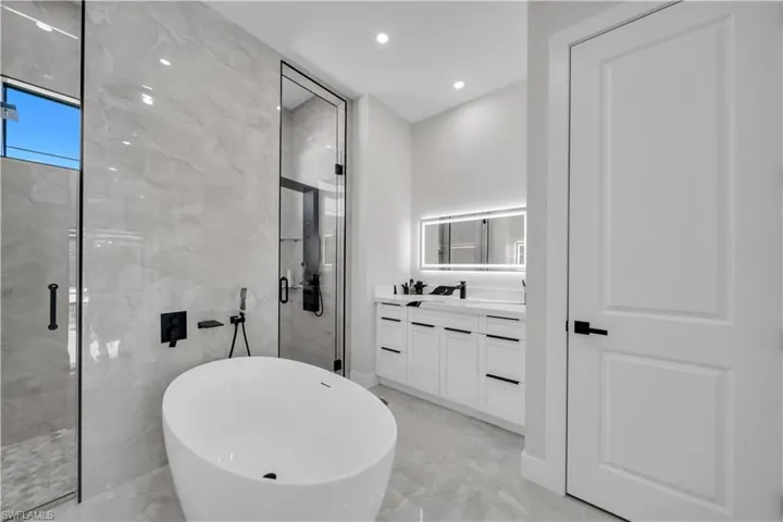 Modern bathroom featuring a freestanding soaking tub, a frameless glass shower enclosure, a dual-sink vanity with white cabinetry, a backlit mirror, and large format tile flooring and shower surround