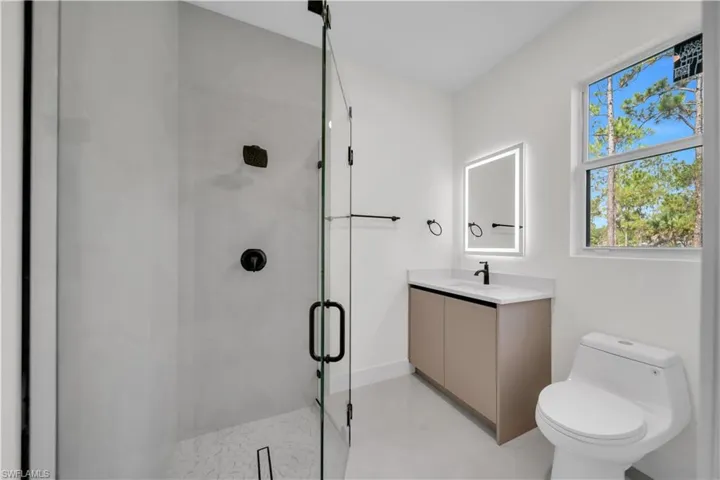 Contemporary third  bathroom featuring a glass-enclosed shower with matte black fixtures, a vanity with a white countertop and integrated sink, a backlit mirror, and a window providing natural illumination