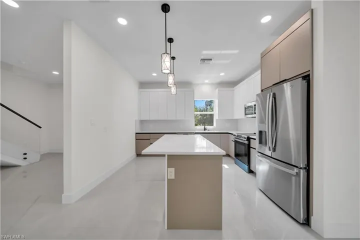 Modern kitchen featuring a central island with a light-toned countertop, white cabinetry, integrated stainless steel appliances, contemporary pendant lighting, and light-colored tile flooring