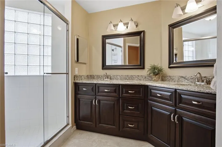 Shower enclosure with glass block window, dark wood-finish vanity with granite-style countertop, dual sinks, and two framed mirrors with vanity lighting