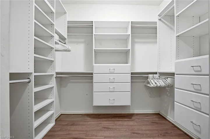 Walk-in closet featuring white built-in shelving, multiple hanging rods, four integrated drawers with silver pulls, and wood-finish flooring