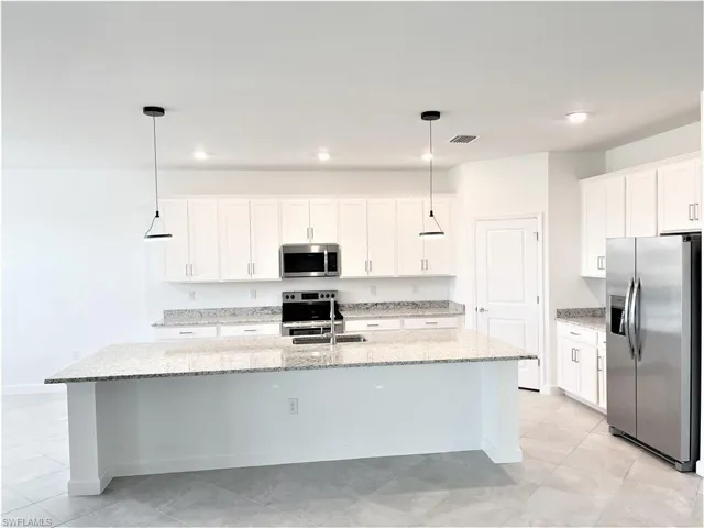 Kitchen with stainless steel appliances, white cabinets, light stone countertops, an island with sink, and pendant lighting