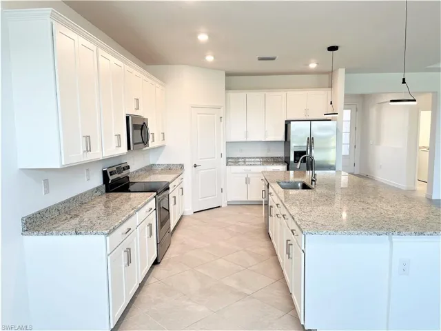 Kitchen with stainless steel appliances, light stone countertops, white cabinets, hanging light fixtures, and an island with sink