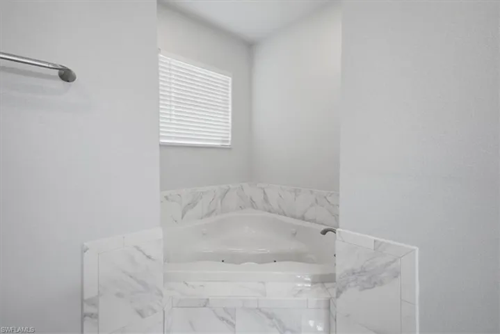 Corner jetted tub featuring a tile surround with marble-look accents, a window with horizontal blinds, and a wall-mounted towel bar