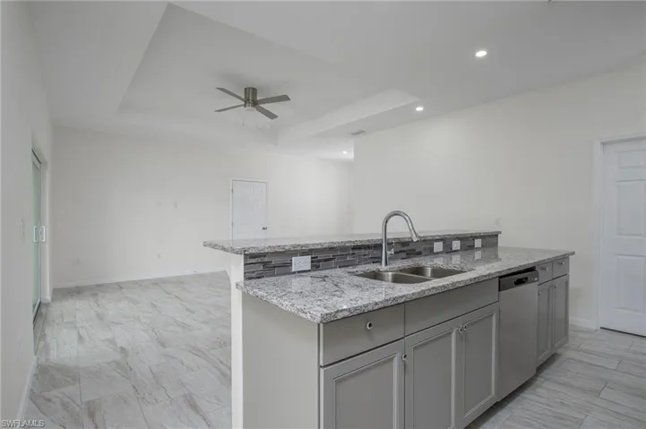 Kitchen with dishwasher, a kitchen island with sink, garbage disposal, ceiling fan, gray cabinetry, and light stone counters with backsplash