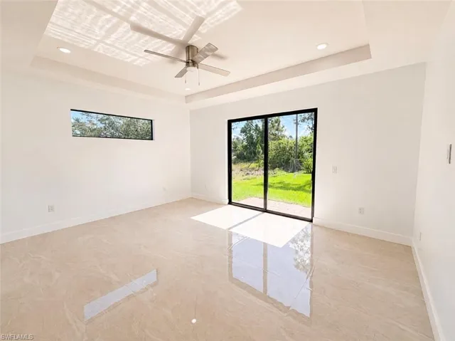 Spacious interior featuring a tray ceiling with recessed lighting, a modern ceiling fan, large format tile flooring, a horizontal window, and sliding glass doors opening to outdoor green space