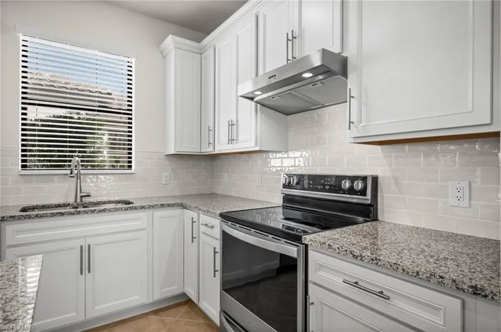 Kitchen with stainless steel electric stove, range hood, white cabinetry, light stone countertops, and light tile patterned flooring