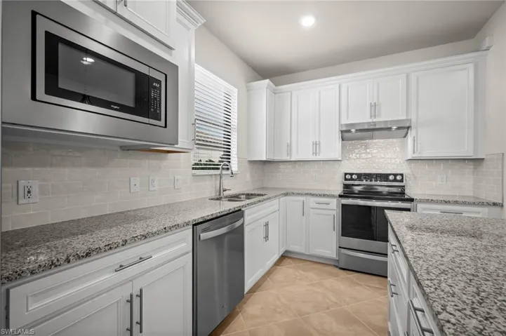Kitchen with stainless steel appliances, white cabinetry, light stone counters, under cabinet range hood, and light tile patterned floors