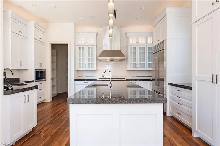 Kitchen featuring wall chimney range hood, white cabinets, built in appliances, decorative light fixtures, and a kitchen island with sink