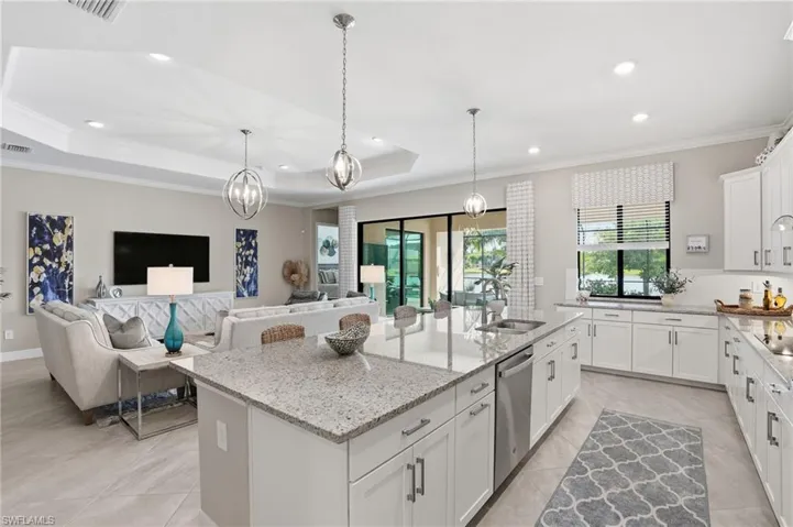 Kitchen with a raised ceiling, white cabinetry, light stone counters, crown molding, and a kitchen island with sink