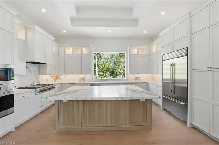 Two tone kitchen featuring a raised ceiling, light stone countertops, stainless steel appliances, two tone cabinetry, and a kitchen island