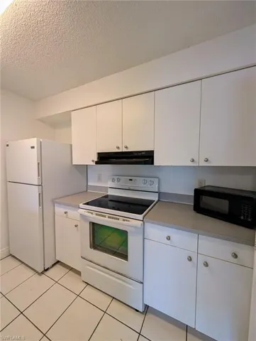 Kitchen with white appliances, white cabinetry, a textured ceiling, light tile patterned floors, and under cabinet range hood
