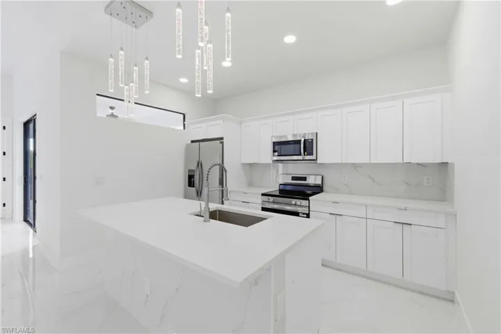 Kitchen featuring stainless steel appliances, white cabinets, a kitchen island with sink, hanging light fixtures, and light marble finish flooring