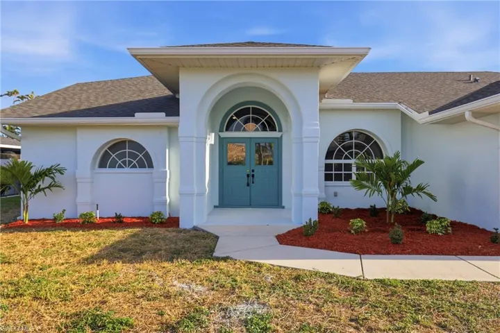 Arched entryway with double teal doors, stucco exterior, half-round windows, and red mulch landscaping beds
