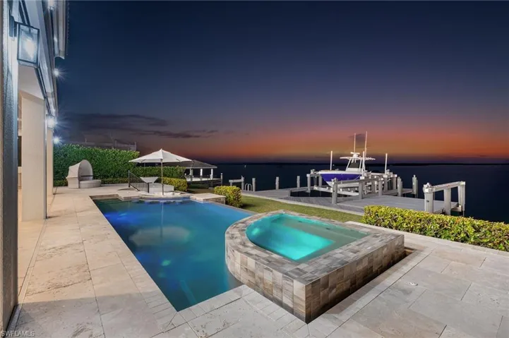 Pool at dusk with an in ground hot tub, a boat dock, a water view, and a patio