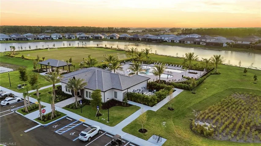 Community clubhouse featuring a tile roof, an adjacent resort-style pool with multiple lounge areas, a covered outdoor pavilion, a large grassy recreation field, and surrounding lush landscaping