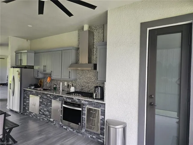 Kitchen featuring backsplash, stone counters, stainless steel appliances, wall chimney exhaust hood, and ceiling fan
