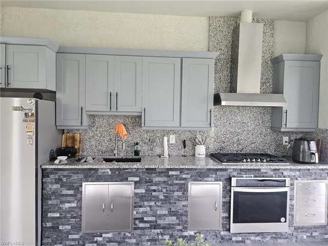 Kitchen featuring backsplash, light stone counters, stainless steel appliances, gray cabinets, and wall chimney exhaust hood