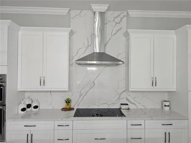 Kitchen featuring black electric cooktop, light stone countertops, wall chimney range hood, and white cabinets