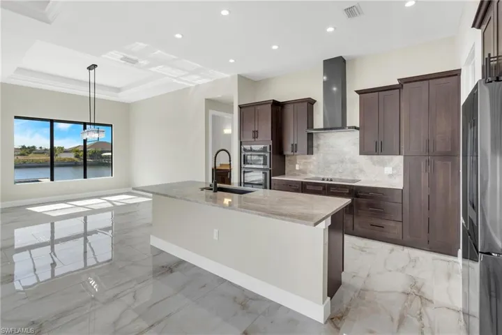 Kitchen with light stone countertops, a kitchen island with sink, stainless steel appliances, light marble finish floors, and range hood