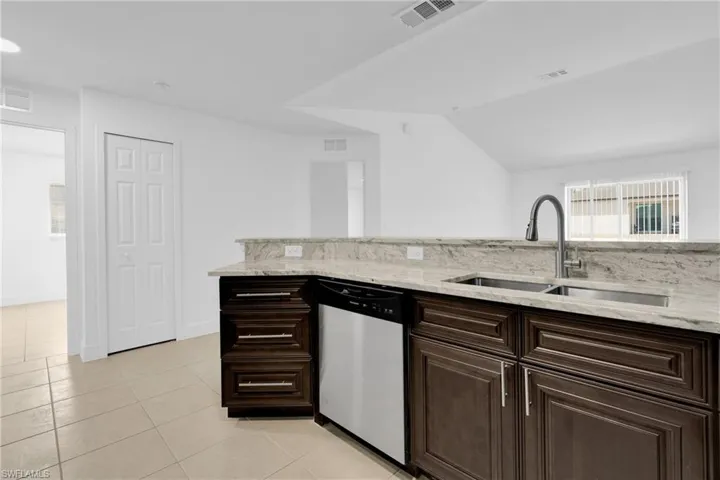 Kitchen featuring dark wood finish cabinetry, stainless steel dishwasher, light stone counters, light tile patterned flooring, and lofted ceiling