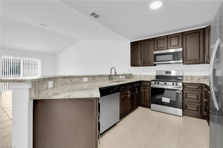 Kitchen with stainless steel appliances, a peninsula, dark wood finish cabinets, light stone counters, and vaulted ceiling