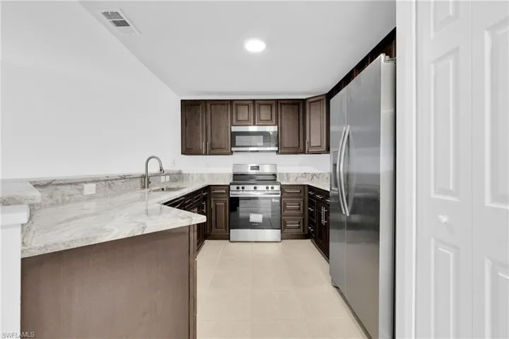 Kitchen featuring stainless steel appliances, dark wood finish cabinetry, light stone countertops, light tile patterned floors, and a peninsula