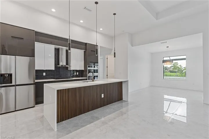Kitchen with hanging light fixtures, decorative backsplash, an island with sink, stainless steel refrigerator with ice dispenser, and wall chimney range hood