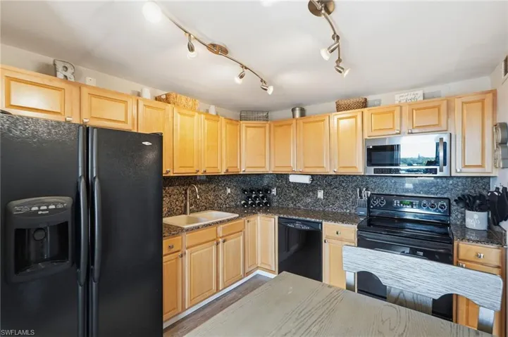 Kitchen with granite counters and backsplash, lighter cabinets, small dining table, black appliances, and a laundry closet.